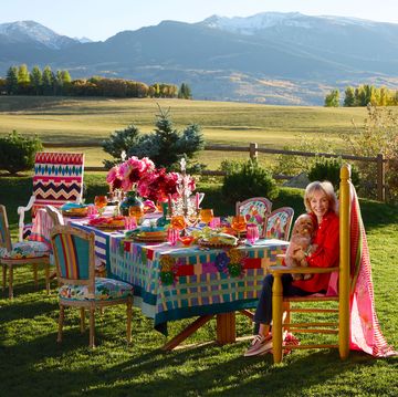 in a garden a woman with a dog on her lap sits at a set table with a bright multicolored tablecloth and bouquets of flowers, surrounded by chairs with bright fabrics, a wood fence, trees, and mountains in the background