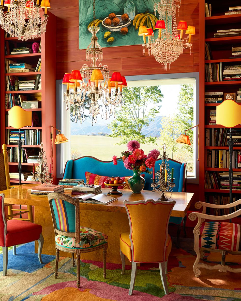 Dining room table with chairs with mismatched cushions and a bright blue sofa, flowers and candelabra on a marble table, paint above the window, sconces and bookshelves on both sides, and three chandeliers