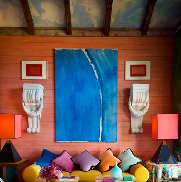 a living room with reddish walls, a mustard sofa with five bright pillows, a large blue painting and other artwork, two end tables with lamps, two cocktail tables, two chairs with striped fabric, turquoise rug