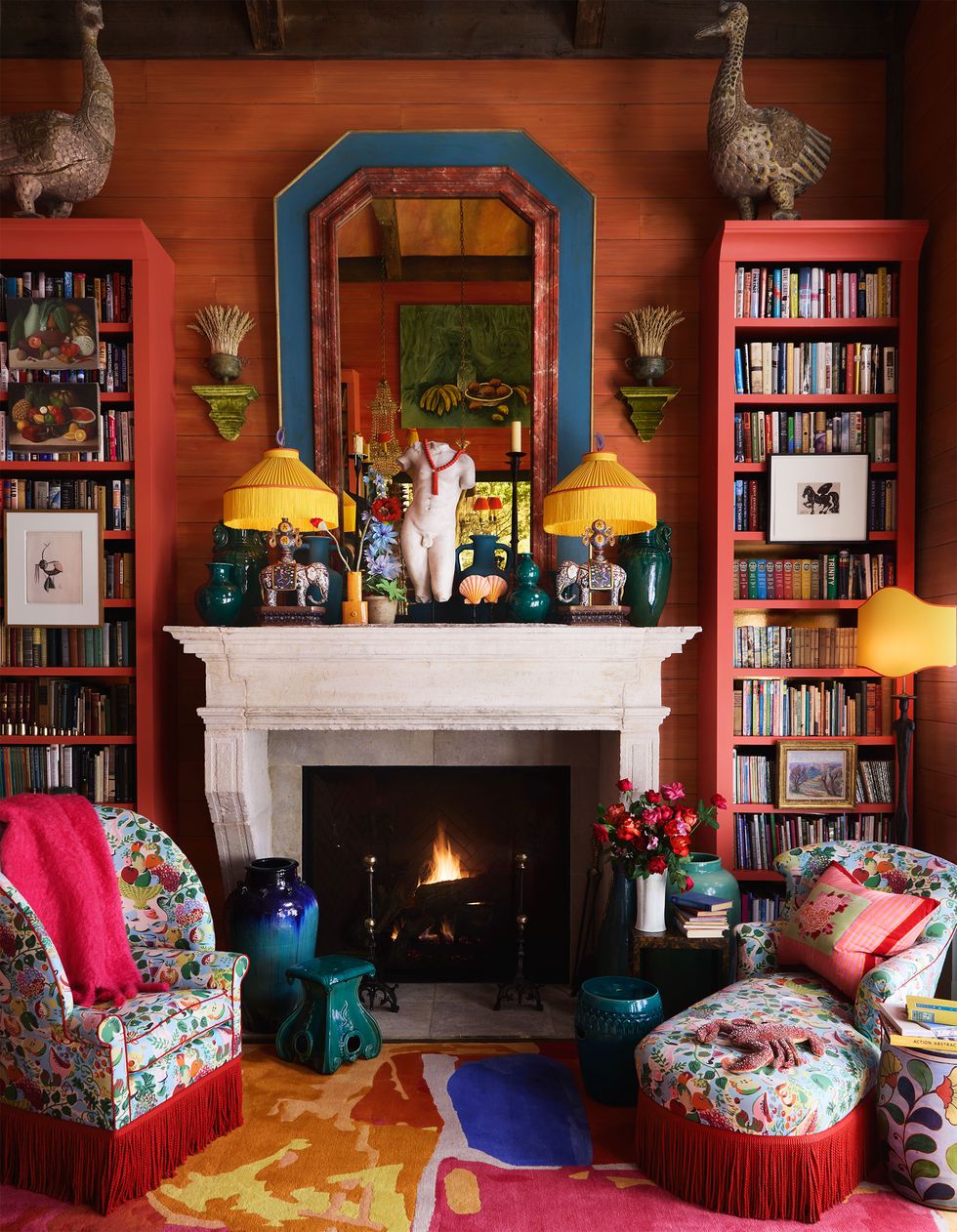 Terracotta colored walls, bookshelves surrounding a fireplace with a mirror, objects and lamps on a mantel, chairs and side tables, a colorful upholstered armchair and chaise longue, a multi-coloured abstract patterned rug