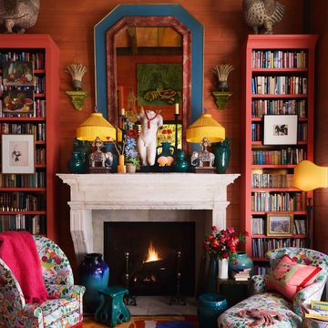 terracotta colored walls, bookshelves flank a fireplace with a mirror, objects, and lamps on mantel, stools and side tables, colorful upholstered armchair and chaise longue, multicolored abstract patterned rug