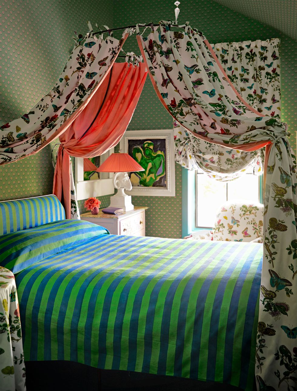 bed with circular canopy in a chintz fabric with butterflies, an armchair and round table in matching fabric, blue and green striped bedcover, nightstand with lamp, green wallpaper with subtle print
