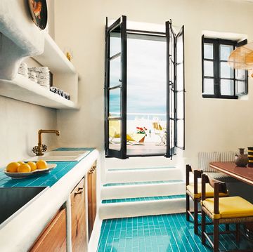 a kitchen with turquoise tiled floor and a wood table with yellow chairs and a banquette with striped fabric, rattan pendant, counter with sink and shelves above and wood cabinets below, steps go to a doorway out to terrace