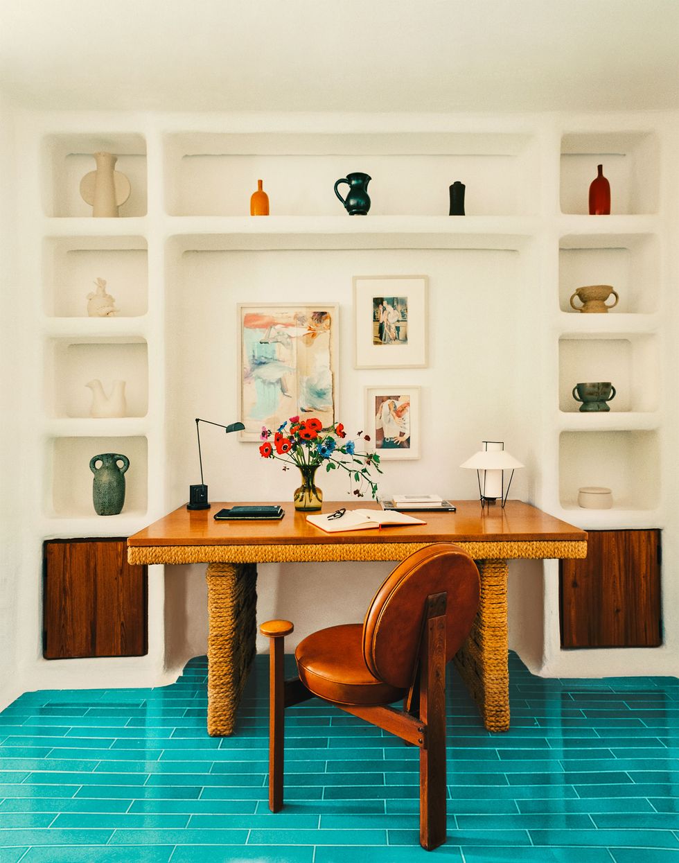 an office area with small artworks above wooden tabletop with legs covered in rattan, wood chair with round leather seat and back, small lamps and a vase on table, inset shelves on either side with objets d'art