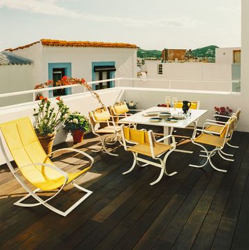 an open terrace with a dark stained deck with yellow back chairs and a white table set with dishes, potted flowering plants, a view of terracotta tiled roofs and mountains in distance