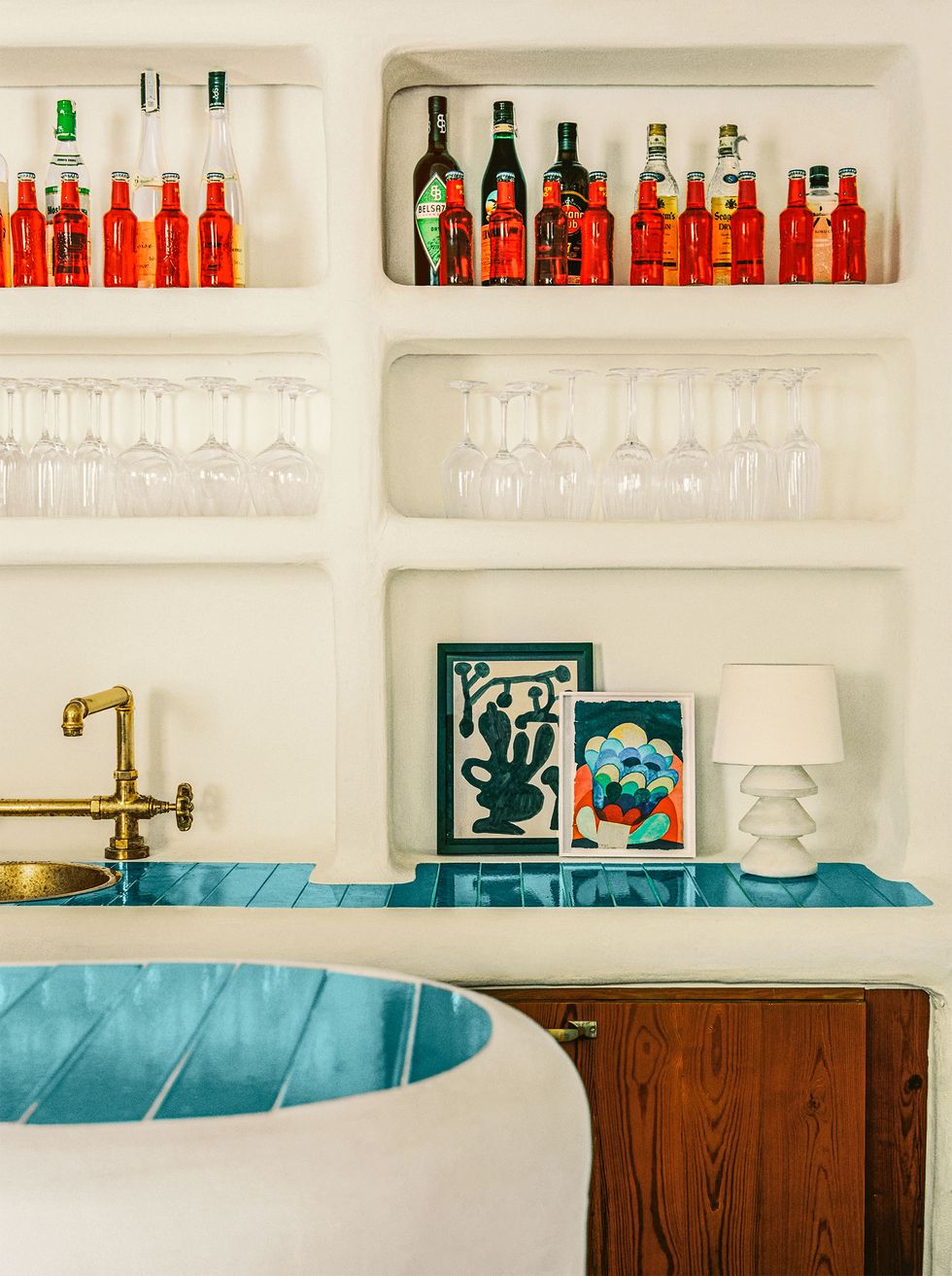 closeup of white stone curved bar, bar and countertop behind it are topped with turquoise tiles, brass faucet and sink, small artworks and a lamp, bottles and glasses are inset into wall above bar