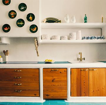 kitchen has turquoise tiled floor, countertop with a sink and stovetop, wood cabinets below and stone shelves above with dishes and decorative objects and a section displaying small plates with fish painted on them