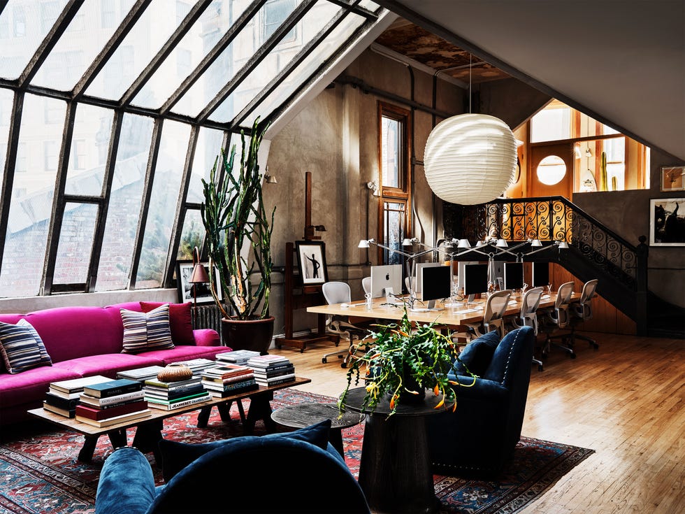 stylish living room with paint tudio windows and a pink sofa and plush blue chair and a black spiral staircase in the back corner and pants and a cocktail table full of books and a long dining table with rolly chairs pulled up to it and a scroll iron staircase leading out in the far end
