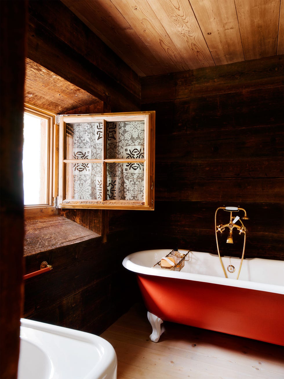 bathroom lined in dark wood, small open, paned window with curtain near a claw foot bathtub with a red exterior, brass fittings, and an attached wire storage rack with soap, white sink and red towel bar, wood ceiling