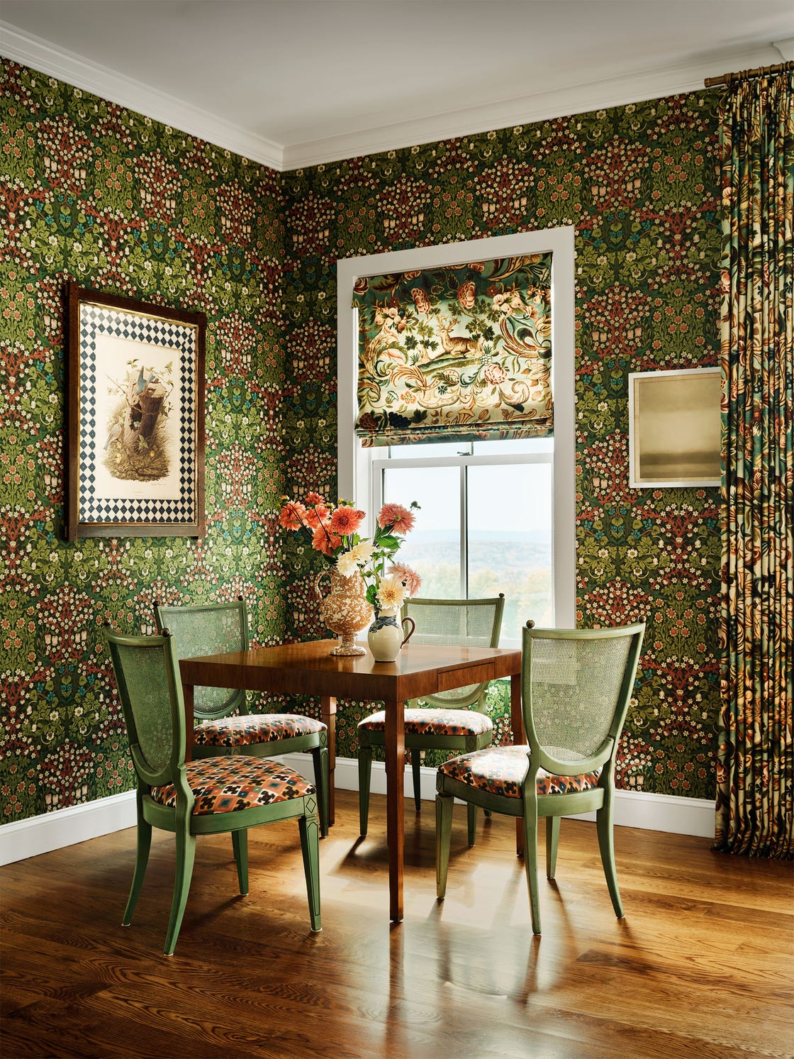 room covered in green based wallpaper, curtains, and window shade with intricate patterns and colors, white trim, wood floor, square wood table and green chairs with patterned seat cushions, flower vases atop table