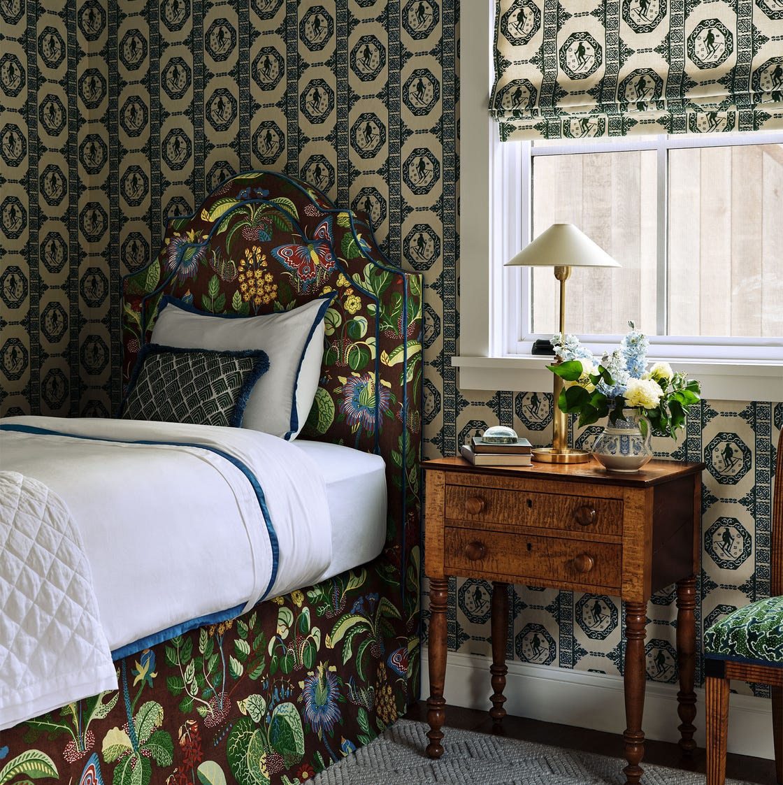 Sheila Bridges - ELLE DECOR bedroom with a green and white patterned design wallpaper and shade, twin bed with a headboard and skirt in a bright pattern with leaves, flowers, and butterflies, wood nightstand with lamp and flower vase