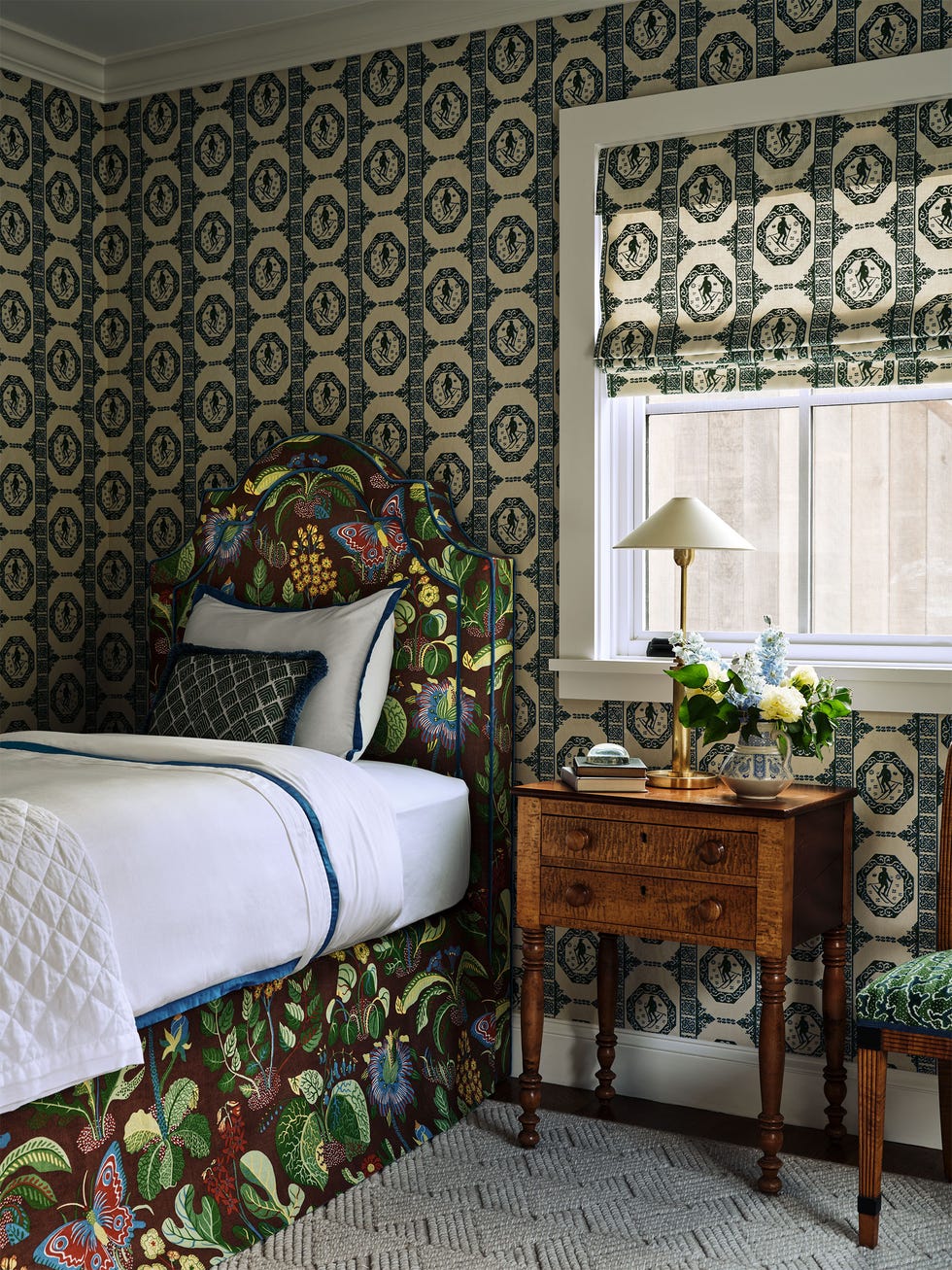 bedroom with a green and white patterned design wallpaper and shade, twin bed with a headboard and skirt in a bright pattern with leaves, flowers, and butterflies, wood nightstand with lamp and flower vase
