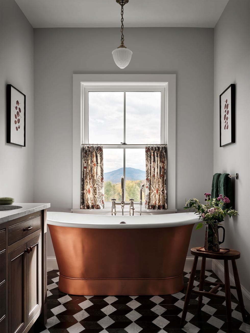 white bathroom with window with a mountain view, half curtains, deep copper tub, side table with vase and flowers, wood cabinet with marble top and sink, black and white diamond floor tiles, pendant over tub