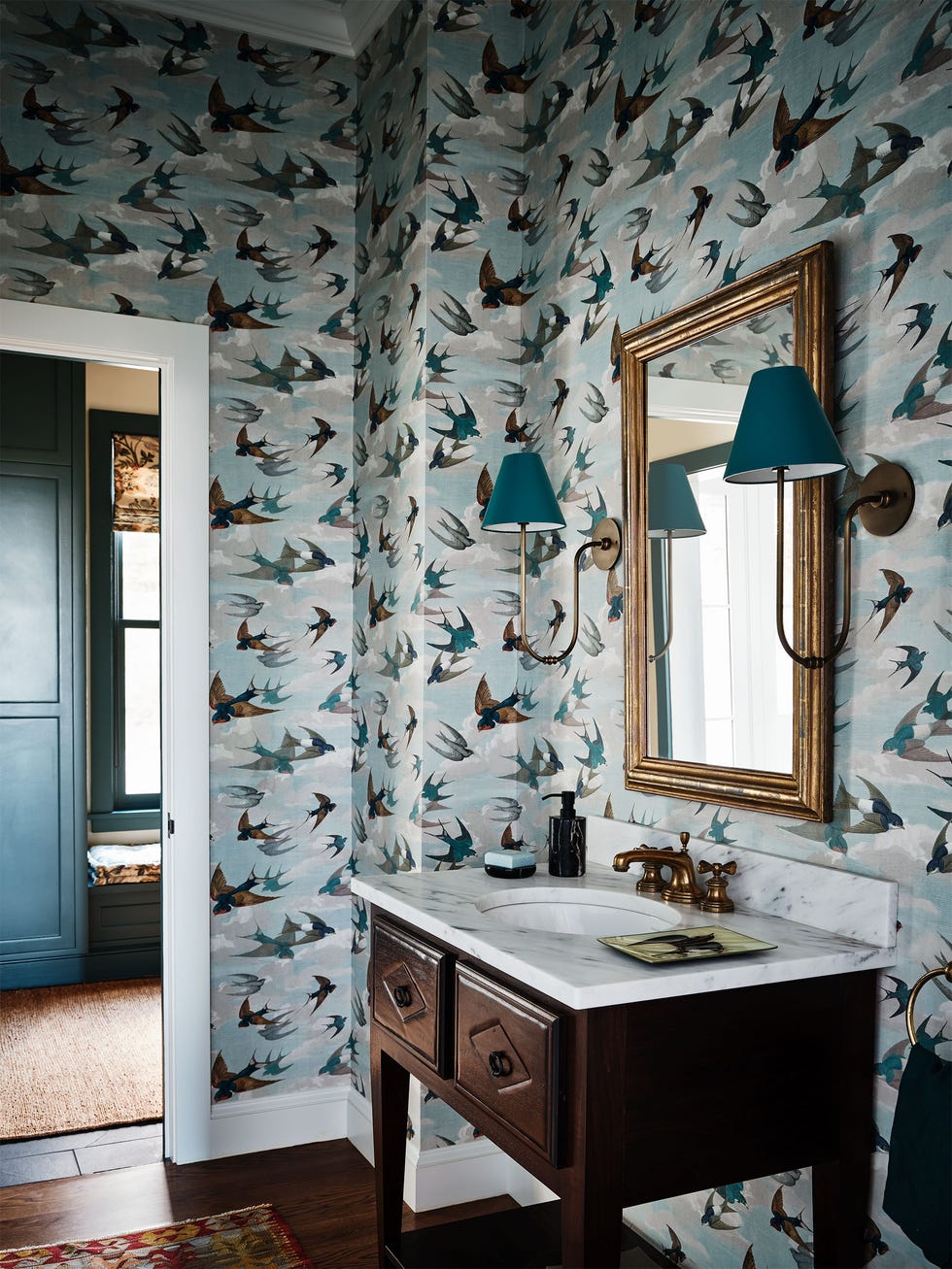 powder room with light blue green wallpaper with clouds and brown, turquoise, and gray birds swooping down, wood base with marble top and sink with bronze fittings, rectangular mirror, sconces with green shades