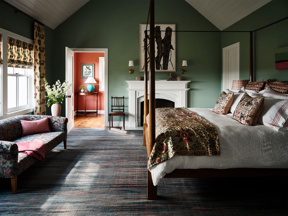bedroom with green walls, white trim, pitched beadboard ceiling, fireplace with decorative mantel, floral sofa in front of windows with roman shades, four poster bed with white bedcove and patterned blanket