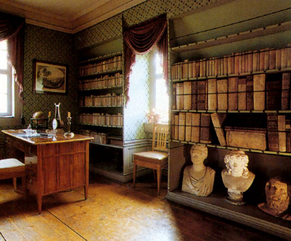 traditional library with green cross hatch and swagged wallpaper and a heavy desk on one side and open wooden shelving holding heavy old bound books and busts