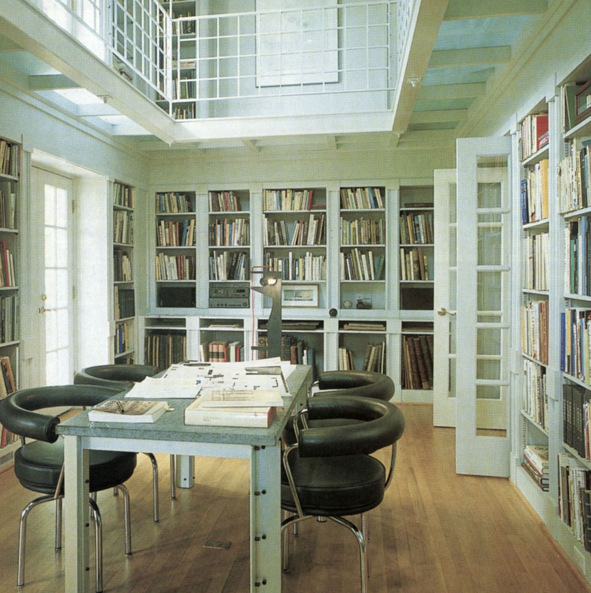 professorial looking white library with open and glassed shelving and a long metal table with leather chairs pulled up to it and a second floor balcony with more shelving