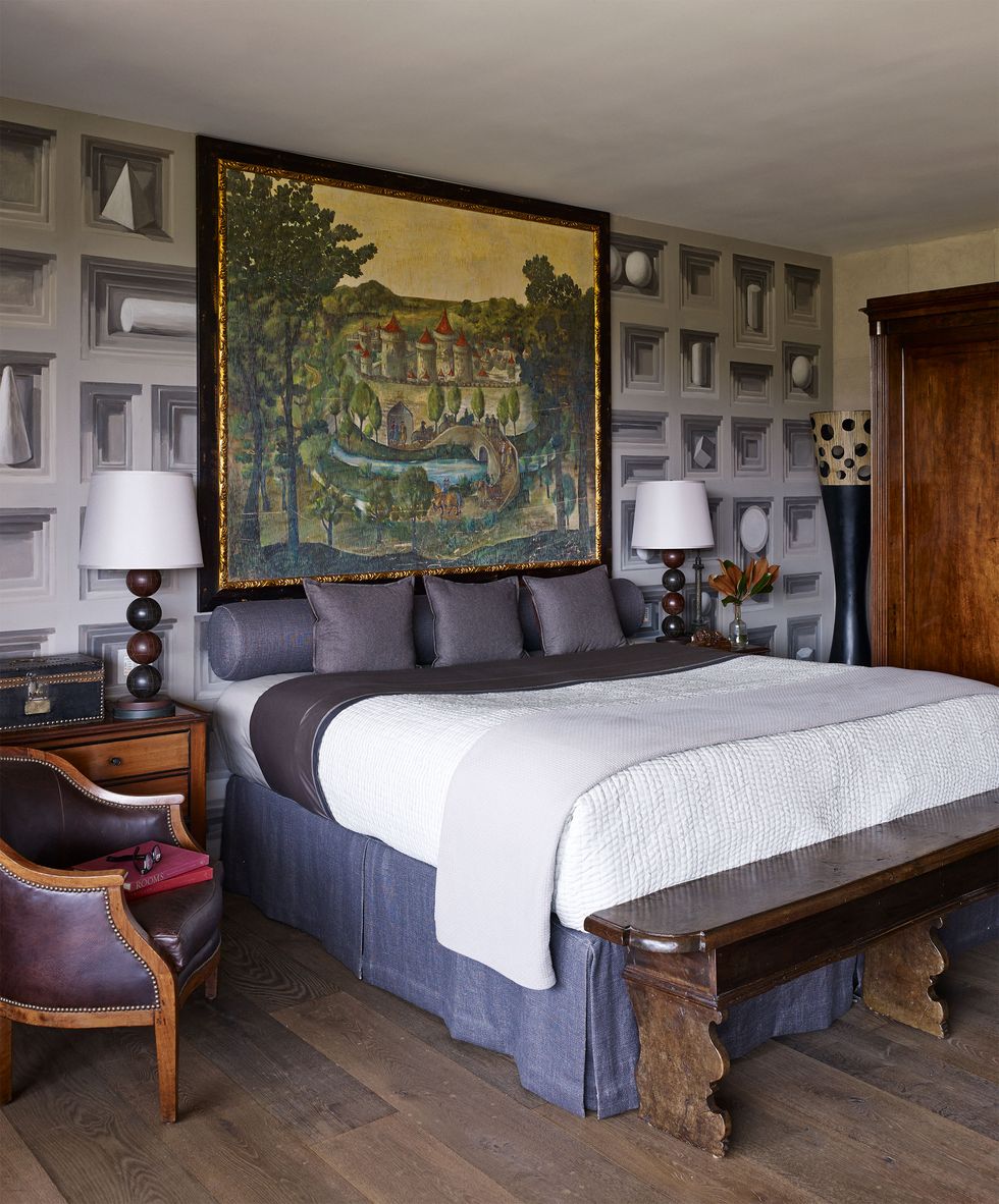 bed with blue and white linens and an old bench at the foot with a large colorful painting over the headboard and the wall is also a large painted mural of boxes and circles in shades of light purplish blue