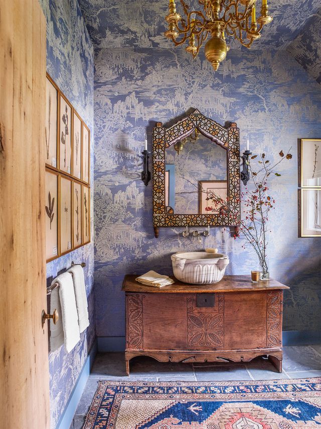 rustic powder room with patterned rug and a distressed wooden console holding a bowl sink and large mosaic mirror above it and tree patterene wallpper on the wall