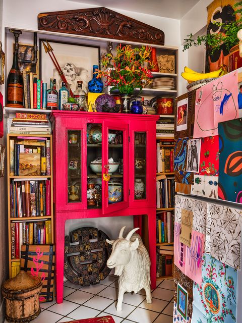 in the kitchen are bookshelves on either side of a hot pink antique pie safe with shelves and art objects on top, a victorian carving above, and a goat sculpture sits beneath it