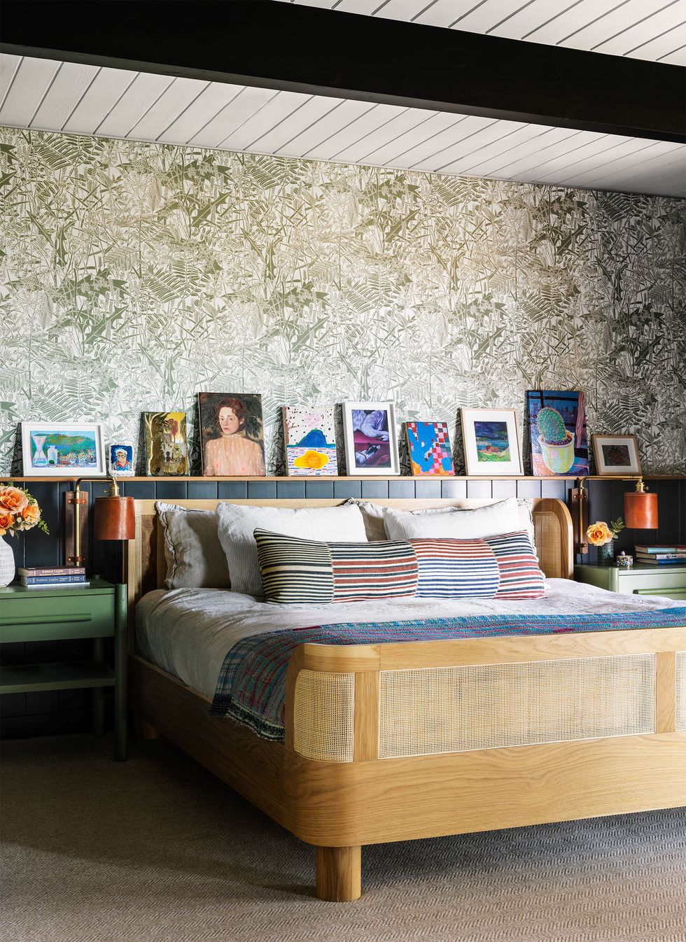 The master bedroom bed has a curved wooden frame with long striped decorative pillows, green nightstands on either side, and artwork on a wainscot shelf against a backdrop of leaf-patterned wallpaper. I am.
