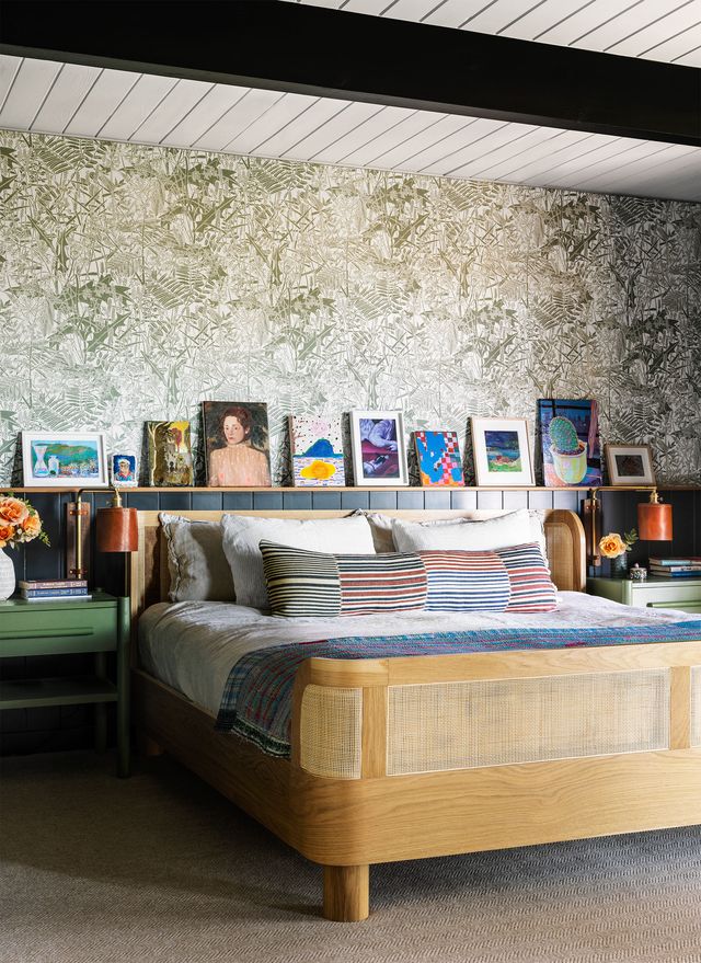 the bed in primary bedroom with curved wood frame and long striped decorative pillow, green nightstands on either side, artworks sit atop wainscoting ledge against leaf patterned wallcovering