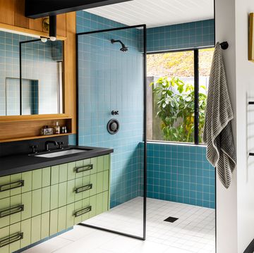 the primary bathroom has a walk in shower with aqua colored tiles, black shower and sink fixtures and countertop with green drawers below and mirror above, deep white bathtub with a sconce and wall sculpture above it