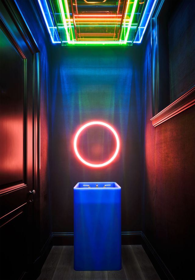 the dark walled powder room has a deep blue sink illuminated by a multicolored florescent ceiling light and a neon lit circular mirror