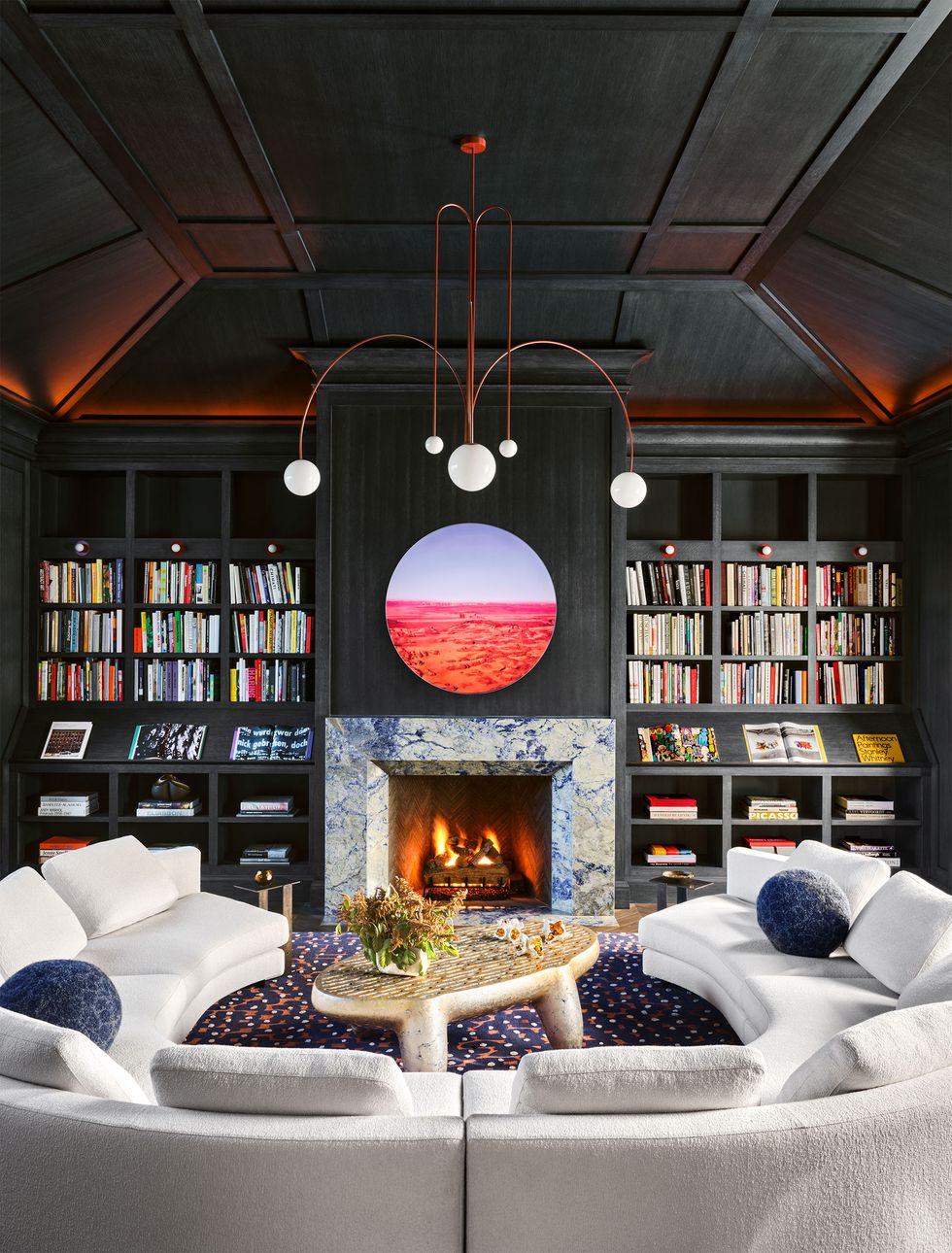 A marble fireplace is flanked by dark wood bookshelves, above which is a circular artwork, a chandelier with five spherical lights, a white circular sofa, and a cocktail table.