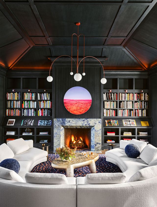 a marble fireplace is flanked by dark wood bookshelves with a circular work of art above it, there is a chandelier with five spherical lights, a white circular sofa, and a cocktail table