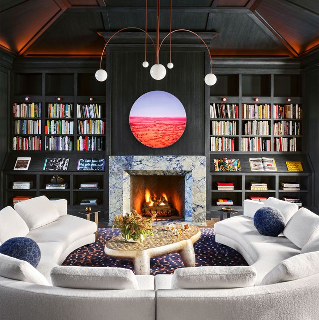 black paneled living area with marble manteled fireplace and adjacent built in shelving on either side and a large circular artwork over the fireplace and a white semicircular sofa on a blue patterned carpet