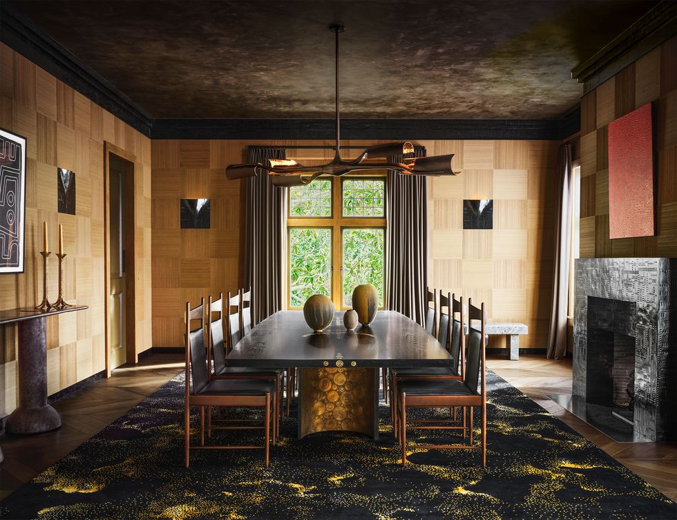 the dining room has wood wall paneling, a curtained floor to ceiling window, a bronze fireplace and dining table with eight chairs, a rug, and wall artworks