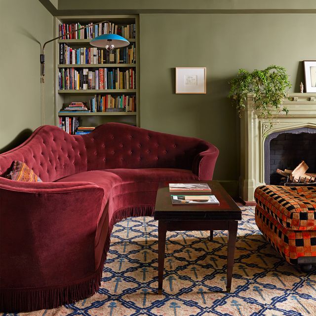 living room with curvy tufted back sofa in warm red velvet and sage green walls and a patterned carpet in blue and pale pink and a wood table and checkered ottoman