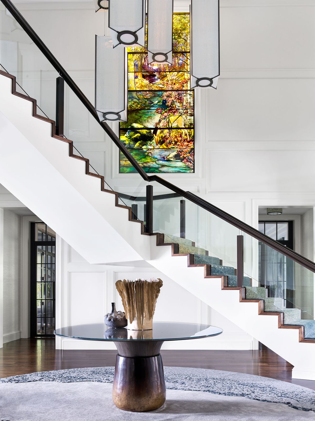 a tiffany studios landscape window overlooks a staircase with glass balustrade and a round glass topped table in foreground
