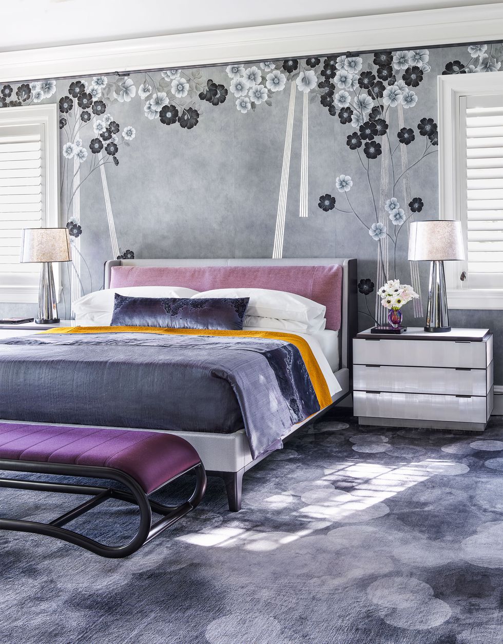 The carpeted master bedroom has floral wallpaper, a bed with a lilac comforter, two nightstands with lamps, and a metal bench with a plum chair.
