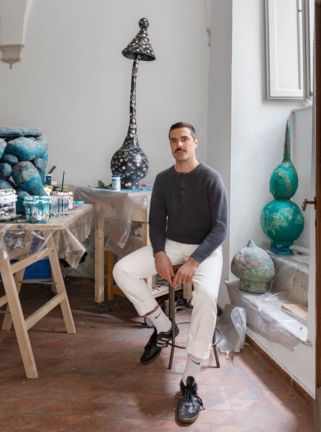 portrait of the artist in his studio seated on a stool with art works behind him