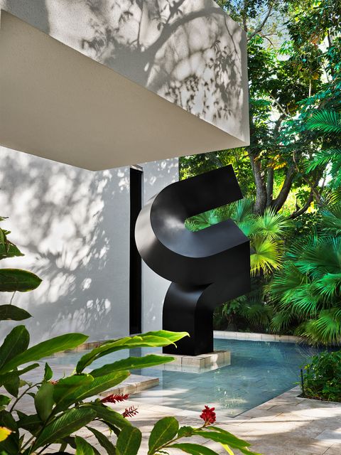 exterior of home with an overhang and a large torquing black steel sculpture anchors a reflecting pool framed by fan palms to the right of the front door