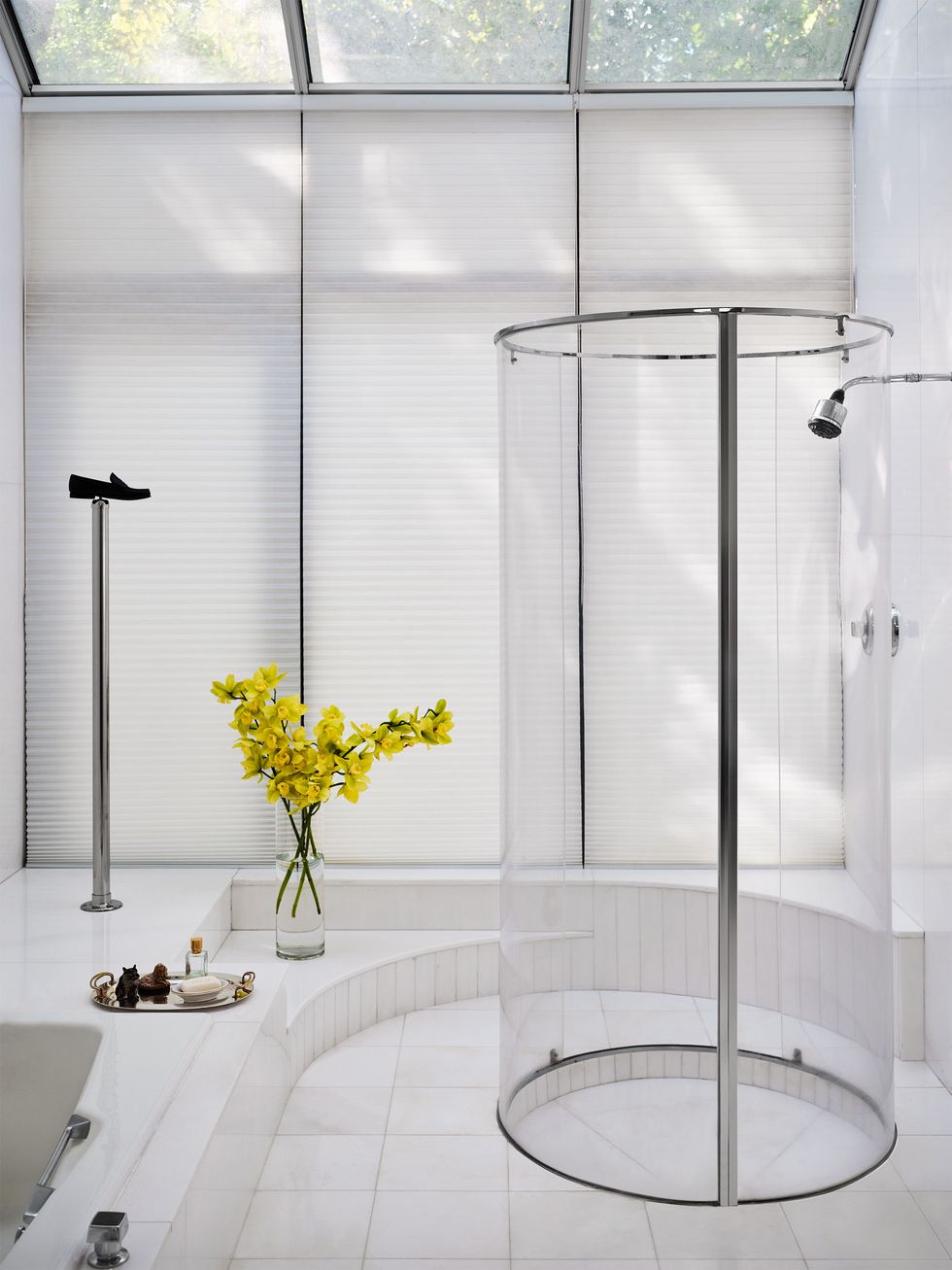 the floor of the primary bathroom is white marble and there is a jacuzzi and a cylindrical shower made of plexiglass, floor to ceiling windows have white honeycomb shades, ceiling windows are uncovered
