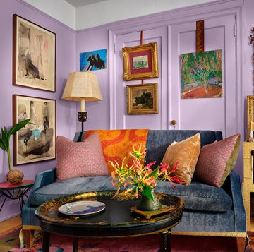 a purple living room with artwork on the walls and a blue velvet covered sofa at center with multiple colored pillows on it and a shelf next to it with books and objects and a large mirror behind it