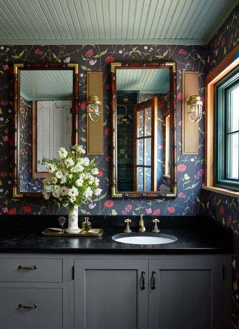 a bathroom has a dark gray vanity with a black marble top and vintage fixtures, a vase with white flowers, wallpaper with a dark gray background and pink flowers and green leaves, twin mirrors, and two ship sconces