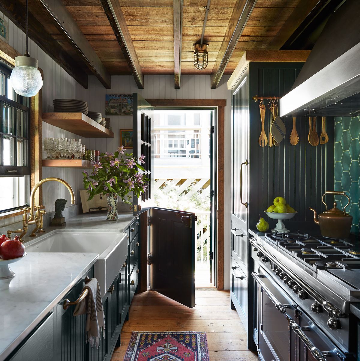 a galley kitchen has cabinets topped with a white marble counter, sink with a glass pendant above, open shelves, an open dutch door, bead board cabinets, stove and oven with tools hanging above, green tiled backsplash