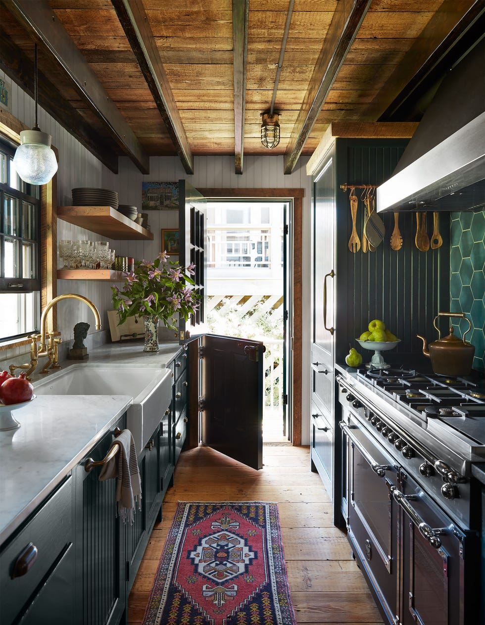 a galley kitchen has cabinets topped with a white marble counter, sink with a glass pendant above, open shelves, an open dutch door, bead board cabinets, stove and oven with tools hanging above, green tiled backsplash