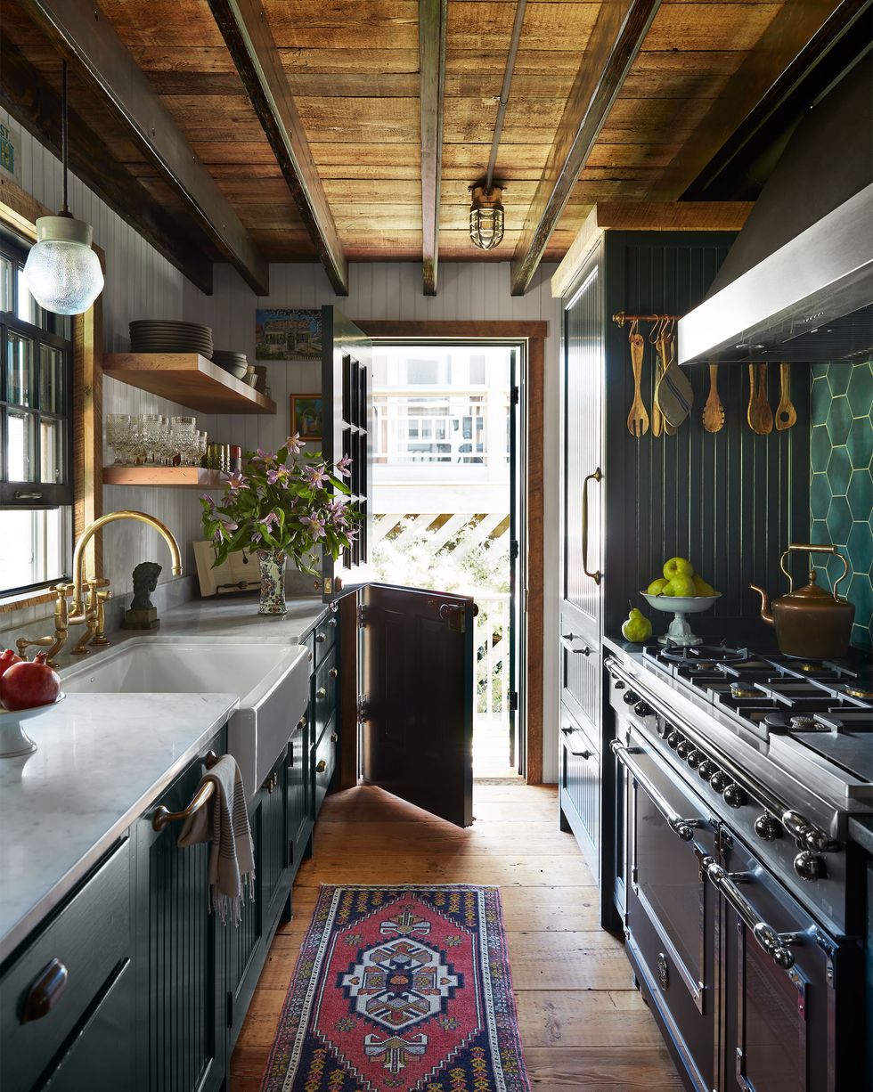 a galley kitchen has cabinets topped with a white marble counter, sink with a glass pendant above, open shelves, an open dutch door, bead board cabinets, stove and oven with tools hanging above, green tiled backsplash