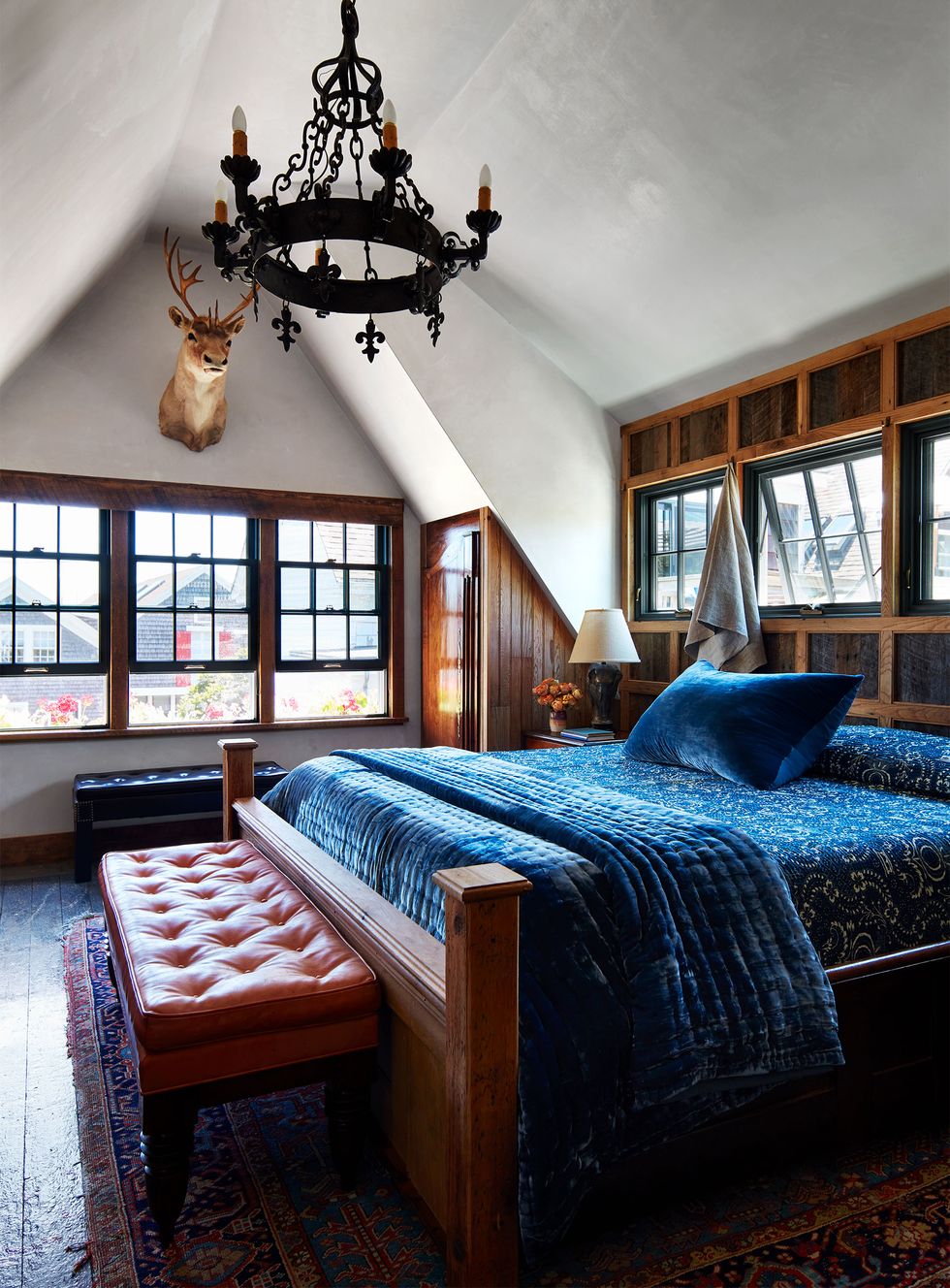 a primary bedroom has deep blue bedding, vented windows above bed, a tufted leather bench at foot of bed and another by a second set of windows, wrought iron chandelier, and a taxidermied deer's head on wall