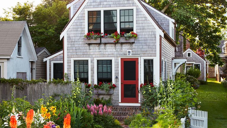 the exterior of a cedar shingled two story cottage with window boxes with red flowers, a red screen door, and brick steps, a white picket fence with roses surrounds tall orange flowers, yellow day lilies, pink foxglove