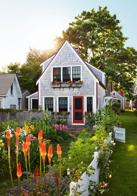 the exterior of a cedar shingled two story cottage with window boxes with red flowers, a red screen door, and brick steps, a white picket fence with roses surrounds tall orange flowers, yellow day lilies, pink foxglove