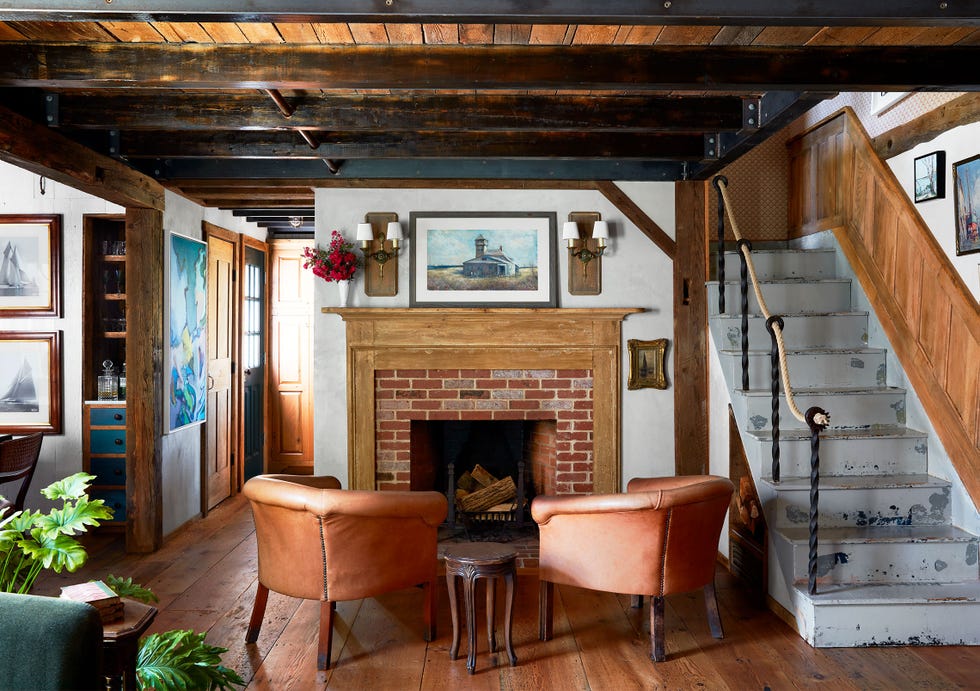 a seating area has a brick fireplace and wood mantel with a painting and sconces above, two leather barrel chairs and small side table, rustic stairs with wrought iron and rope banister, wood floor and exposed beam ceiling