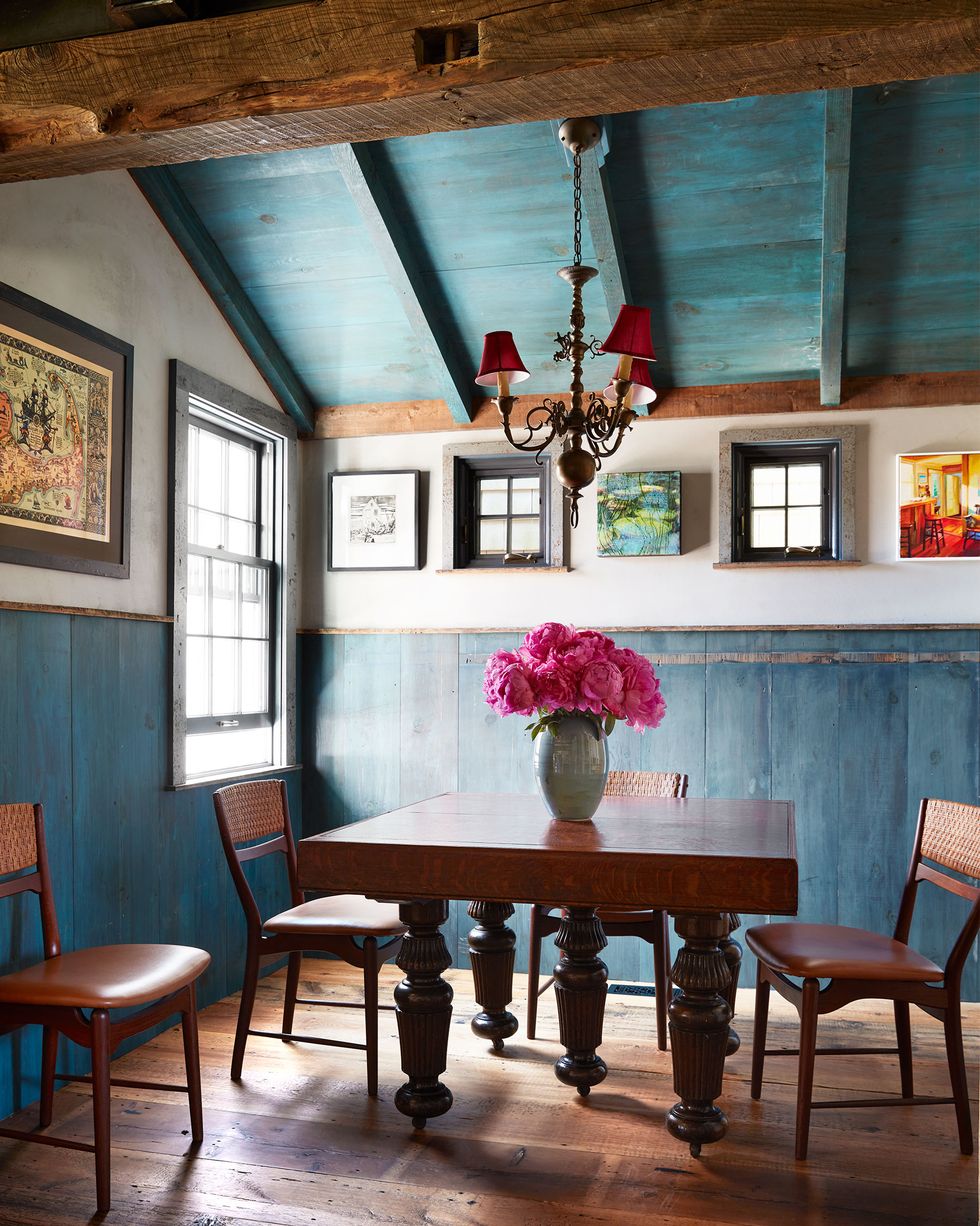 in a dining room is an edwardian wood table with deep pink flowers in a vase, four midcentury danish chairs, wainscoting in original blueberry stain, three armed chandelier with shades, vintage cape cod map on wall