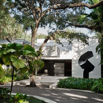 exterior of the modern home with a circular drive, a black steel sculpture, an oak tree in front of steps leading to a black set in entrance, with trees and shrubs surrounding the property
