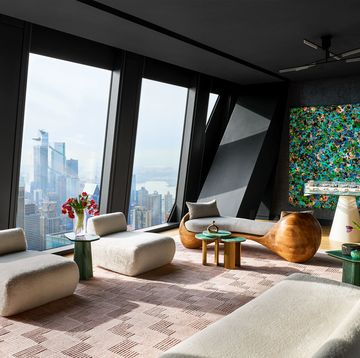 a penthouse living room has floor to ceiling windows, an armless sofa and chairs in an off white textured fabric, geometric rug, a curved wood chaise with fabric cushion, foosball table, large colorful painting